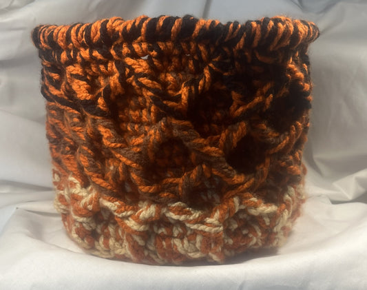 A handmade basket made from chunky, textured yarn in a variegated pattern of orange, dark brown, and cream, set against a draped white fabric background.